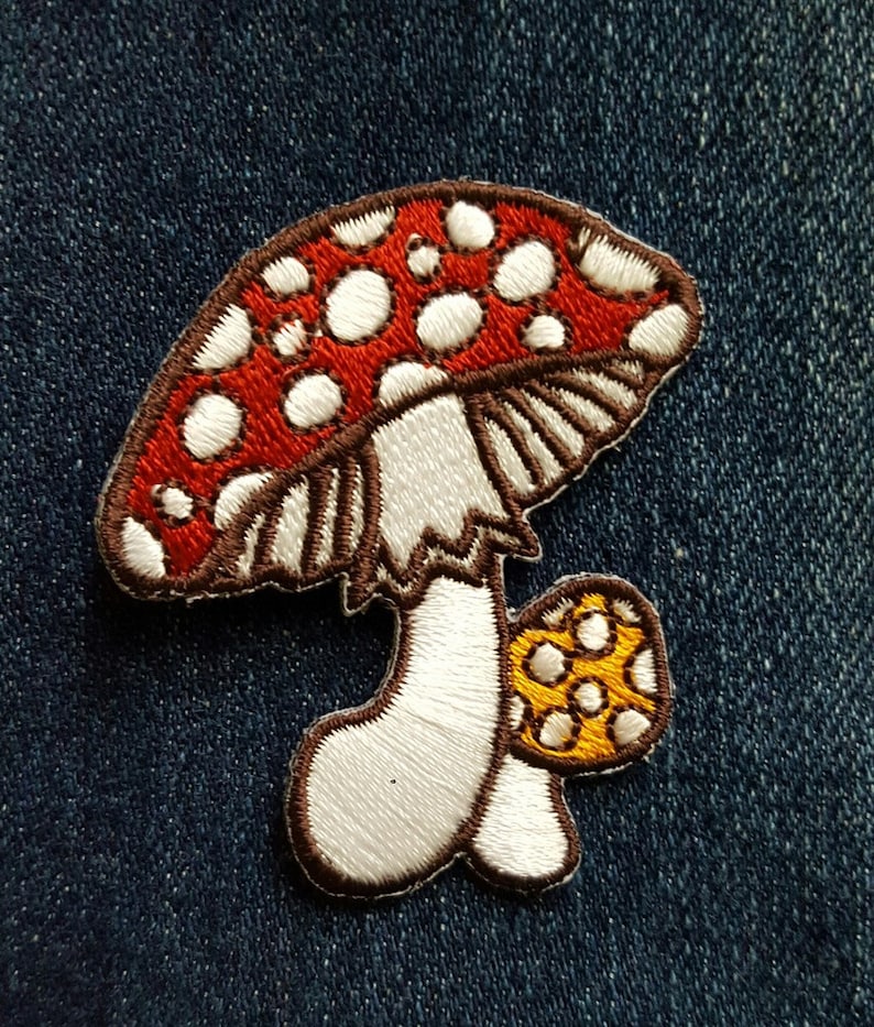 K&ouml;nnte beinhalten: Gestickter Pilz-Aufn&auml;her mit rotem Hut und wei&szlig;en Punkten, wei&szlig;em Stiel und einem kleineren gelben Pilz. Der Aufn&auml;her hat eine braune Umrandung und befindet sich auf einem blauen Jeansstoff. Ideal zum Aufn&auml;hen auf Kleidung.