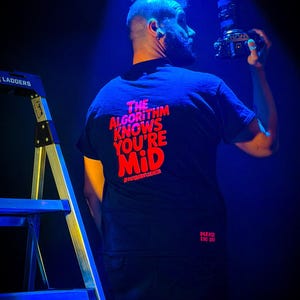 May include: A black t-shirt featuring red text on the back that reads THE ALGORITHM KNOWS YOU'RE MiD and #NOTANINFLUENCER, with the text PLEASE LOG OFF printed in smaller red letters near the bottom hem of the shirt.
