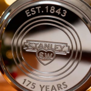 May include: Close-up of a shiny, silver-colored metal emblem. The emblem features the text "EST. 1843" at the top, with "STANLEY S.W." in a heart-shaped design in the center. The words "175 YEARS" are at the bottom.