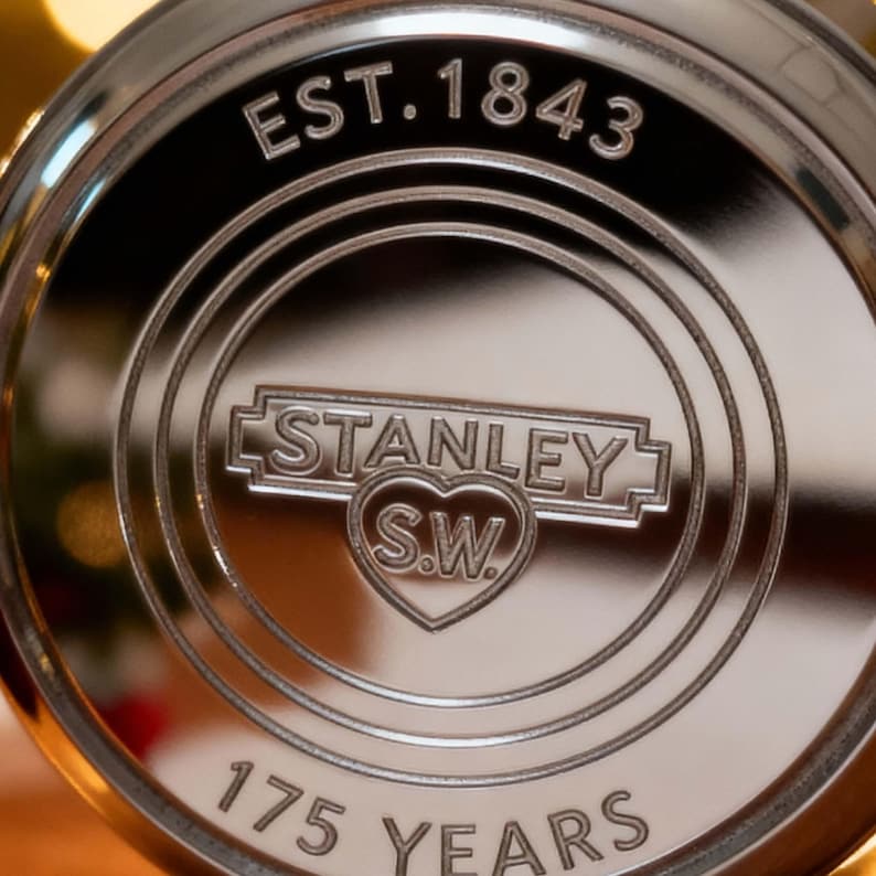 May include: Close-up of a shiny, silver-colored metal emblem. The emblem features the text "EST. 1843" at the top, with "STANLEY S.W." in a heart-shaped design in the center. The words "175 YEARS" are at the bottom.