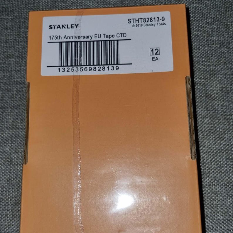 May include: A Stanley 175th Anniversary EU Tape CTD box. The box is orange with white text and a barcode. The text includes the Stanley logo, product information, and a serial number. The box contains 12 units.