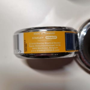 May include: Close-up of a Stanley tape measure with a black and yellow label. The label reads "MADE IN THAIKAND" and includes the Stanley logo and website address. The tape measure is partially visible.