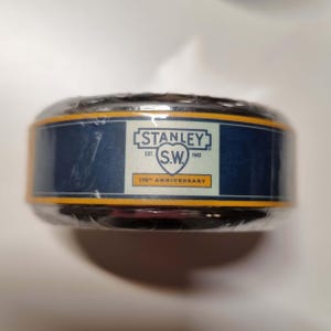 May include: A cylindrical container wrapped in clear plastic. The container has a navy blue band with gold trim and the text "STANLEY S.W. EST. 1843" and "175th ANNIVERSARY" in white.