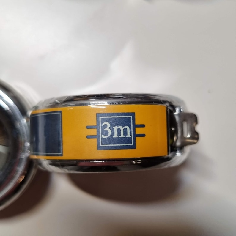 May include: A close-up of a chrome tape measure with a yellow and navy blue label. The label displays the text "3m" in a white box, indicating a measurement of 3 meters, or approximately 9.8 feet. The tape measure is partially visible.