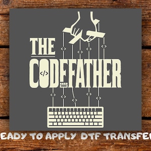 May include: A dark gray square DTF transfer featuring the off-white text THE CODEFATHER. The letter O is replaced by code brackets. Above the text is a hand operating marionette strings connected to a computer keyboard at the bottom.