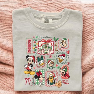 May include: A light gray Comfort Colors sweatshirt featuring a colorful Christmas-themed graphic. The graphic includes various Disney characters like Mickey, Minnie, Donald, and Chip and Dale, along with the text "Merry Christmas".