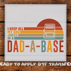 May include: White square with the text "I KEEP ALL MY DAD JOKES IN A DAD-A-BASE" in a retro design. The design includes a sunset with a memory card icon. The words "READY TO APPLY DTF TRANSFER" are at the bottom.