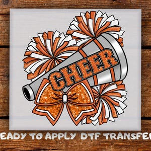 May include: A square design featuring a silver megaphone with the word "CHEER" in orange glitter lettering. Orange and white pom-poms and a bow are also included in the design. The words "READY TO APPLY DTF TRANSFER" are at the bottom.
