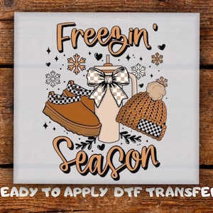 May include: A square graphic with the words "Freezin' Season" in orange script. The image features a brown boot, a tumbler with a bow, and a knit hat with a pom-pom. Snowflakes and hearts are scattered around the objects.