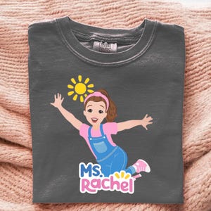 May include: Dark gray t-shirt featuring a cartoon illustration of a girl with a pink shirt, blue overalls, and a yellow sun. The text "MS. Rachel" is below the illustration. The shirt is made of a soft material.
