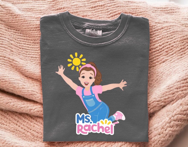 May include: Dark gray t-shirt featuring a cartoon illustration of a girl with a pink shirt, blue overalls, and a yellow sun. The text "MS. Rachel" is below the illustration. The shirt is made of a soft material.