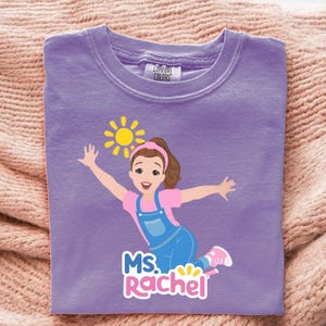 May include: A lilac Comfort Colors t-shirt featuring a cartoon illustration of a girl with outstretched arms, a sun, and the text "MS. Rachel". The girl wears a pink shirt, blue overalls, and pink shoes.