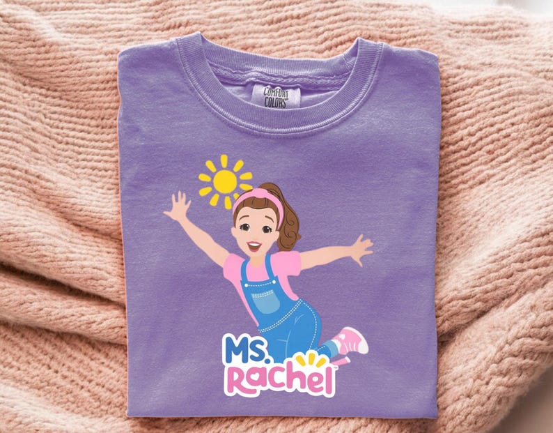 May include: A lilac Comfort Colors t-shirt featuring a cartoon illustration of a girl with outstretched arms, a sun, and the text "MS. Rachel". The girl wears a pink shirt, blue overalls, and pink shoes.