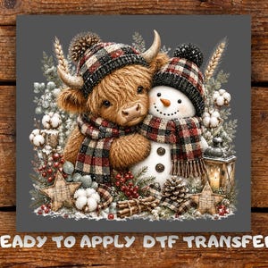 May include: A DTF transfer featuring a brown Highland cow and a white snowman wearing matching red, black, and white plaid hats and scarves. The scene includes cotton bolls, red berries, pine cones, wooden stars, and a lantern on a gray background.
