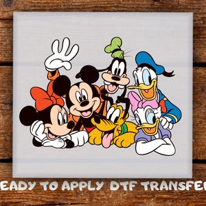 May include: A vibrant graphic featuring Mickey Mouse, Minnie Mouse, Goofy, Donald Duck, Daisy Duck, and Pluto. The characters are in a group pose, with the text "READY TO APPLY DTF TRANSFER" at the bottom. The image is on a light grey background.