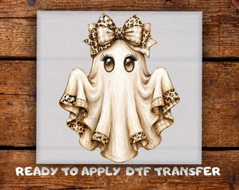 May include: A graphic illustration of a ghost wearing a large leopard print bow on its head. The ghost has large brown eyes with long eyelashes and a beige sheet body with leopard print trim at the bottom edges. Text reads: READY TO APPLY DTF TRANSFER.