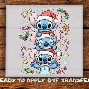 May include: A light gray DTF transfer sheet featuring a stack of three blue alien characters wearing red and white Santa hats. The design includes festive holiday elements like candy canes, a gingerbread man, holly, stars, and snowflakes.