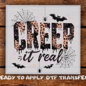 May include: A square graphic transfer featuring the text CREEP it real in a black and peach leopard print pattern with a dripping effect. The design includes black spiderwebs, two spiders, and three bats on a white background with orange splatters.