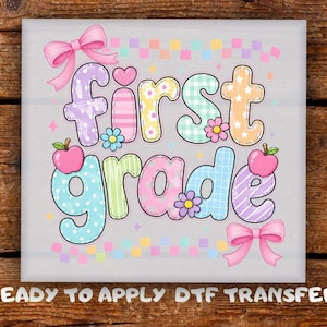 May include: A DTF transfer featuring the text first grade in colorful, patterned bubble letters. The design includes two pink bows, two red apples, and small flowers in shades of purple, blue, and yellow, with a checkered border at the top and bottom.