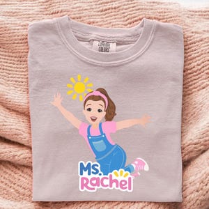 May include: A light purple Comfort Colors sweatshirt featuring a cartoon illustration of a woman with brown hair, a pink shirt, and blue overalls. The design includes a yellow sun and the text "MS. Rachel".