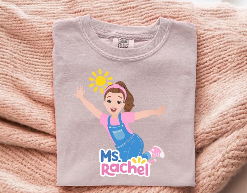May include: A light purple Comfort Colors sweatshirt featuring a cartoon illustration of a woman with brown hair, a pink shirt, and blue overalls. The design includes a yellow sun and the text "MS. Rachel".
