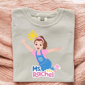 May include: A light gray sweatshirt featuring a cartoon illustration of a woman with brown hair, a pink shirt, and blue overalls. The design includes a yellow sun and the text "MS. RACHEL" in pink and blue.