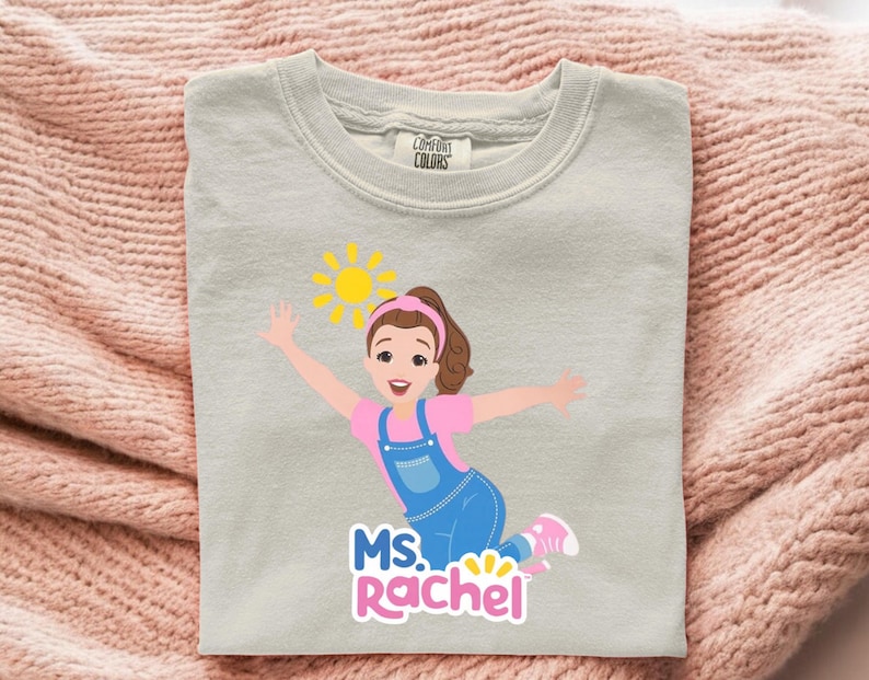 May include: A light gray sweatshirt featuring a cartoon illustration of a woman with brown hair, a pink shirt, and blue overalls. The design includes a yellow sun and the text "MS. RACHEL" in pink and blue.