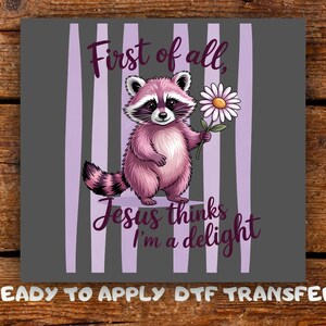 May include: A pink raccoon illustration holding a daisy, set against a grey background with vertical lavender stripes. Text reads "First of all, Jesus thinks I'm a delight." The words "READY TO APPLY DTF TRANSFER" are at the bottom.