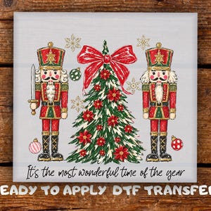 May include: A DTF transfer featuring two nutcrackers in red and green uniforms flanking a Christmas tree decorated with red poinsettias and a large red bow. Gold snowflakes and ornaments surround the figures with the text It's the most wonderful time of the year and READY TO APPLY DTF TRANSFER.