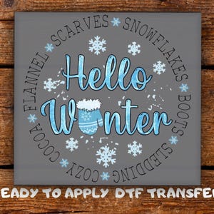May include: A gray square with the words "Hello Winter" in blue script, surrounded by snowflakes and winter-themed words. The words "Ready to Apply DTF Transfer" are at the bottom.