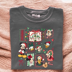May include: Dark gray crew neck t-shirt featuring a colorful Christmas design. The design includes Mickey Mouse, Minnie Mouse, Donald Duck, and other Disney characters in festive attire. The word "Christmas" is also visible.