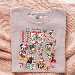 May include: A light purple Comfort Colors sweatshirt featuring a festive Christmas design. The design includes various Disney characters like Mickey, Minnie, Donald, and Goofy, all wearing Santa hats. The words "Merry Christmas" are also included in the design.