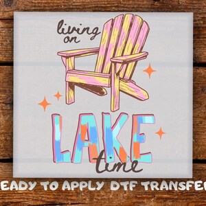 May include: A square graphic transfer featuring a pink and yellow Adirondack chair with the text living on LAKE time in brown and multi-colored brushstroke lettering, accented by four orange four-pointed stars on a light gray background.