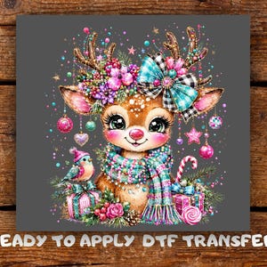 May include: A DTF transfer featuring a brown fawn with large eyes, wearing a teal and pink striped scarf and a plaid bow between its antlers. The design includes colorful ornaments, a small bird, gift boxes, and candy, with the text READY TO APPLY DTF TRANSFER.