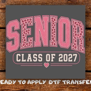 May include: A grey rectangular DTF transfer featuring the word SENIOR in large pink letters with a leopard print pattern. Below, the text CLASS OF 2027 appears in white within a pink outlined box, accompanied by a small pink heart icon centred below.