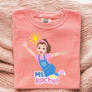 May include: A coral-colored sweatshirt featuring a graphic of a cartoon girl with a sun and the text "MS. Rachel". The girl is wearing a pink shirt, blue overalls, and pink shoes. The sweatshirt is made of a soft material.