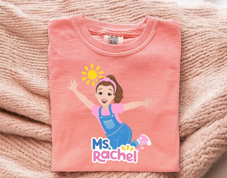 May include: A coral-colored sweatshirt featuring a graphic of a cartoon girl with a sun and the text "MS. Rachel". The girl is wearing a pink shirt, blue overalls, and pink shoes. The sweatshirt is made of a soft material.