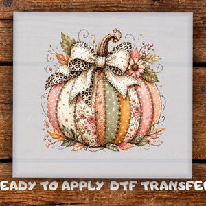 May include: A square DTF transfer featuring a pumpkin with vertical panels in shades of pink, green, and orange, decorated with floral and polka dot patterns. A large cream-colored bow with a black leopard print sits on top of the pumpkin.