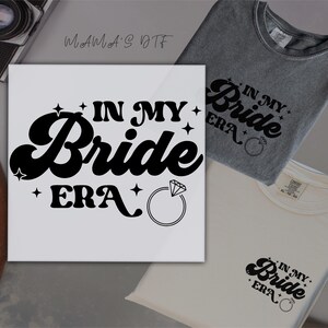 May include: A white square sign and two t-shirts with the text "IN MY Bride ERA" in black. The sign features a diamond ring graphic. One t-shirt is grey, and the other is white.