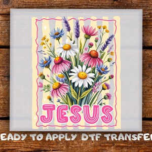 May include: A rectangular DTF transfer sheet featuring a vibrant bouquet of daisies, coneflowers, and lavender against a pale yellow and white striped background. The word JESUS is printed in large, pink, rounded letters at the bottom.