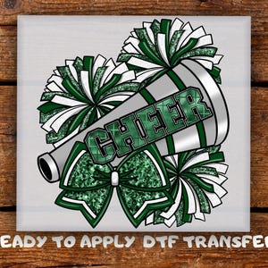 May include: A cheerleading-themed design featuring a megaphone with the word "CHEER" in green glitter. Green and white pom-poms and a bow surround the megaphone. The text "READY TO APPLY DTF TRANSFER" is at the bottom.