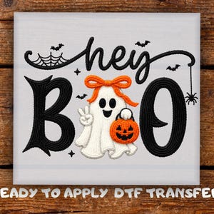 May include: An embroidered design featuring the black text hey BOO with a white ghost wearing an orange bow, holding an orange jack-o'-lantern bucket, and making a peace sign. The design includes black spiderwebs, bats, and a small spider.