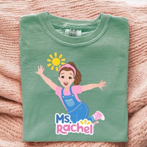 May include: A sage green t-shirt featuring a cartoon illustration of a girl with brown hair, wearing a pink shirt and blue overalls, with a yellow sun graphic. The text "MS. RACHEL" is below the illustration.