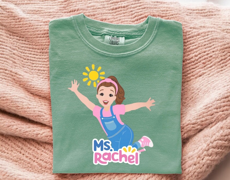 May include: A sage green t-shirt featuring a cartoon illustration of a girl with brown hair, wearing a pink shirt and blue overalls, with a yellow sun graphic. The text "MS. RACHEL" is below the illustration.