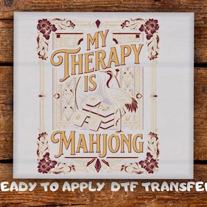 May include: A square DTF transfer featuring the text MY THERAPY IS MAHJONG in gold and dark red. The design includes a crane illustration, a stack of mahjong tiles, and ornate floral borders in a vintage art deco style on a white background.