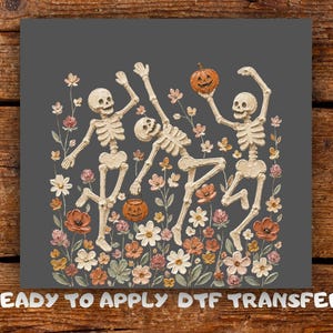 May include: A DTF transfer featuring three off-white skeletons dancing among a field of colourful flowers in shades of orange, pink, and white. One skeleton holds an orange jack-o'-lantern, and another carries a small pumpkin bucket. Text reads READY TO APPLY DTF TRANSFER.