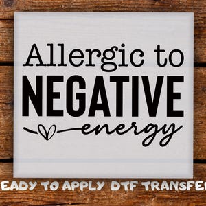 May include: A white square with the words "Allergic to NEGATIVE energy" in black, with a heart symbol. The phrase "READY TO APPLY DTF TRANSFER" is at the bottom. The background is a wooden surface.