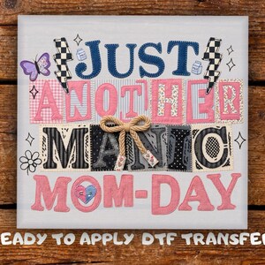 May include: A white fabric square with the text "JUST ANOTHER MANIC MOM-DAY" in various colors and fonts. The design includes a butterfly, lightning bolts, and a bow. The words are in a patchwork style.