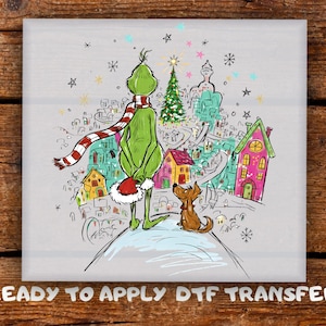 May include: A colourful illustration of a green character with a red and white striped scarf and a Santa hat, overlooking a whimsical town. A small brown dog sits nearby. The text "READY TO APPLY DTF TRANSFER" is at the bottom.