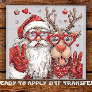 May include: A DTF transfer featuring a cartoon illustration of Santa Claus and a reindeer wearing red heart-shaped glasses. Both figures are making peace signs with their hands. The design includes red hearts, snowflakes, and the text READY TO APPLY DTF TRANSFER.
